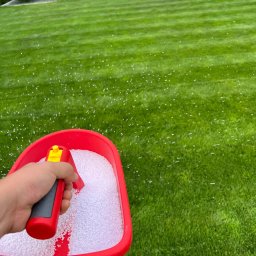 Jarbud - Ręczne rozsiewanie białego nawozu na soczyście zielonym trawniku z widocznymi pasami koszenia, w tle dekoracyjne krzewy i meble ogrodowe.