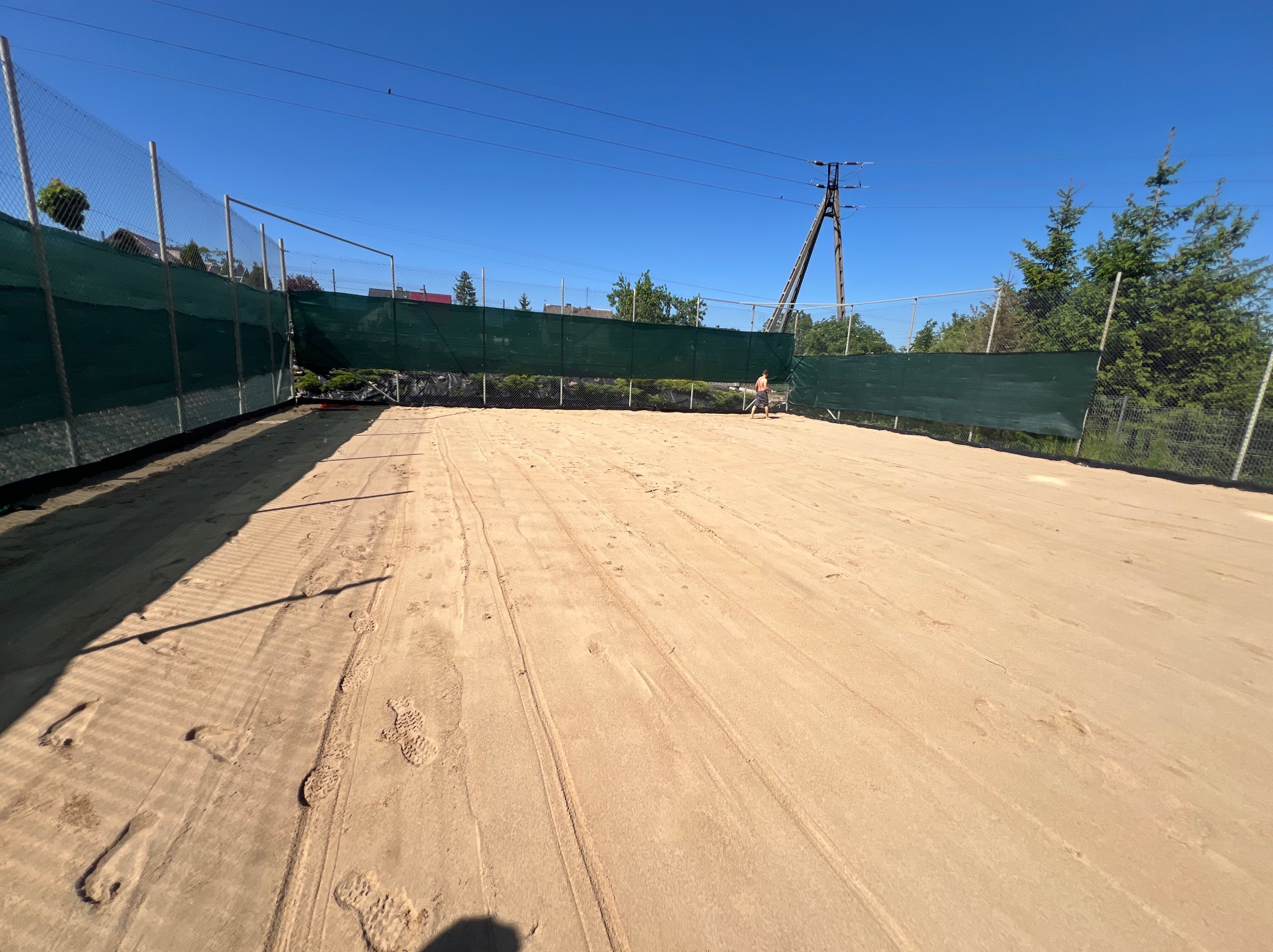 Teren przygotowany pod boisko do siatkówki plażowej, otoczony siatką z zielonym płótnem, widoczne ślady stóp i opon, w tle słup wysokiego napięcia i drzewa, słoneczny dzień.