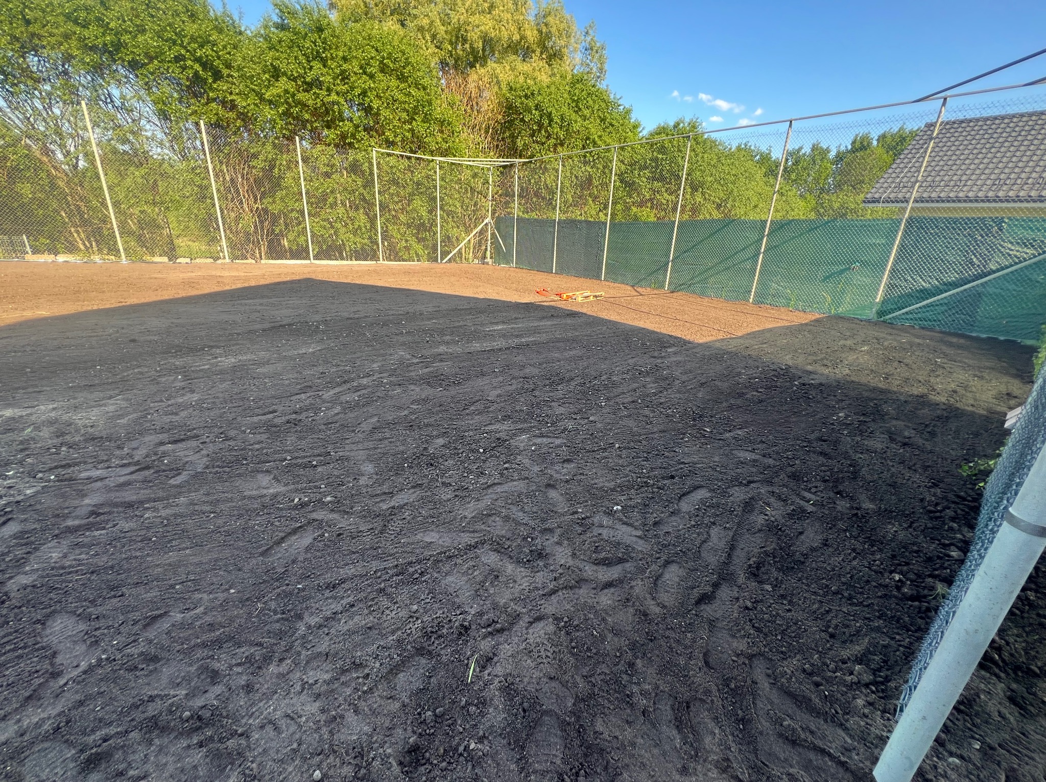 Przygotowany teren pod trawnik z widocznymi śladami opon, ogrodzony siatką z zieloną plandeką w tle, z narzędziami w oddali.