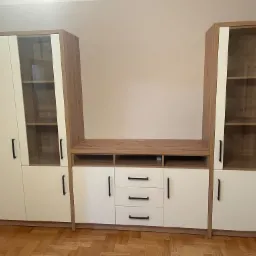 Zestaw mebli pokojowych z jasnego drewna i białych frontów, składający się z dwóch wysokich szaf z przeszklonymi witrynami i szafek dolnych z szufladami, na parkiecie.