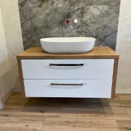 Nowoczesna łazienka z drewnopodobną podłogą i wiszącą szafką z blatem drewnianym, na której stoi owalna, biała umywalka nablatowa; na ścianie widoczne przyłącza wodne.