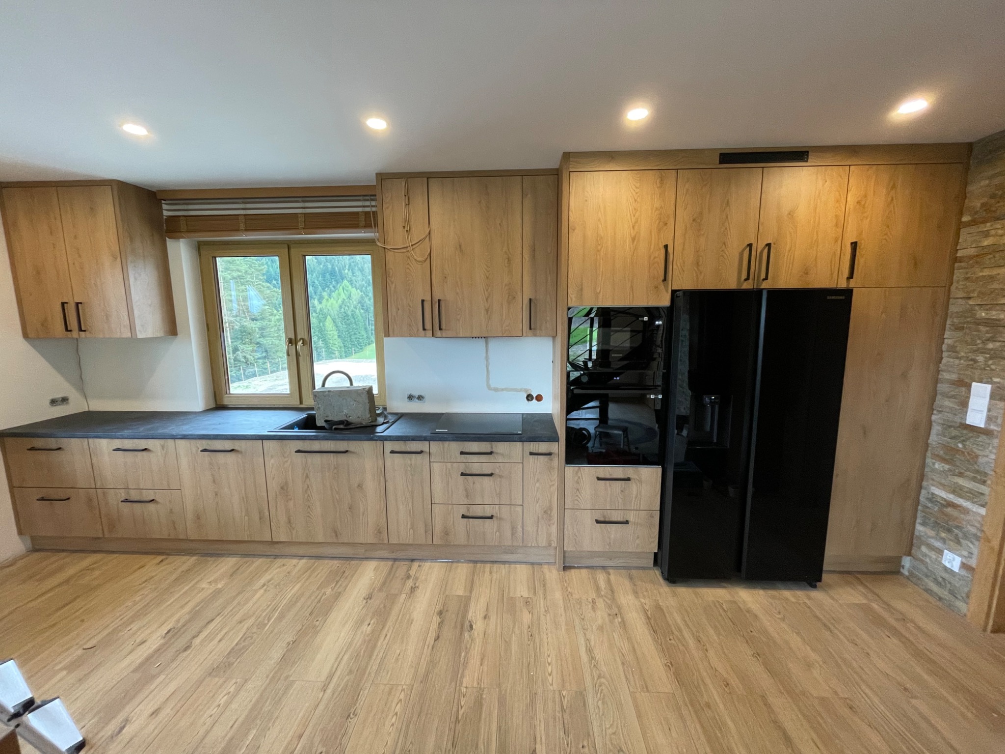 Jasna, drewnopodobna zabudowa kuchenna z czarnymi blatami i sprzętami AGD, widok na zielony krajobraz za oknem, częściowo zakryty zlew z betonowym obciążnikiem.