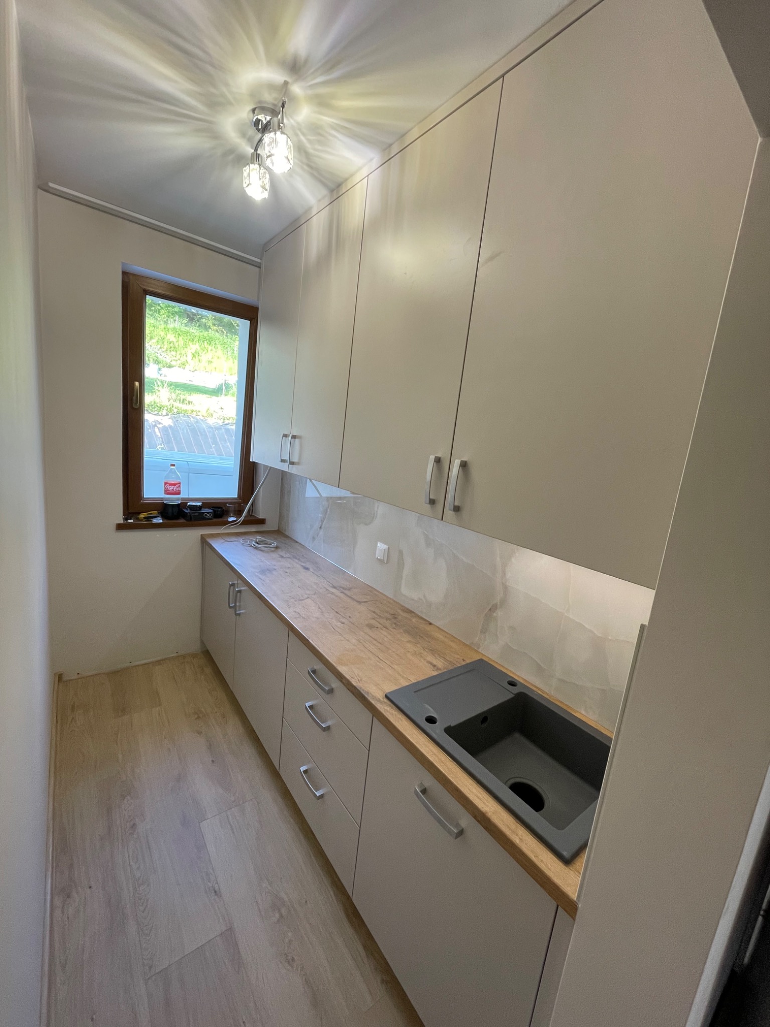 Wąska kuchnia z beżowymi szafkami wiszącymi i dolnymi, drewnianym blatem, szarym zlewozmywakiem i oknem z widokiem na zieleń. Na parapecie butelka Coca-Coli i akcesoria. Oświetlenie punktowe...