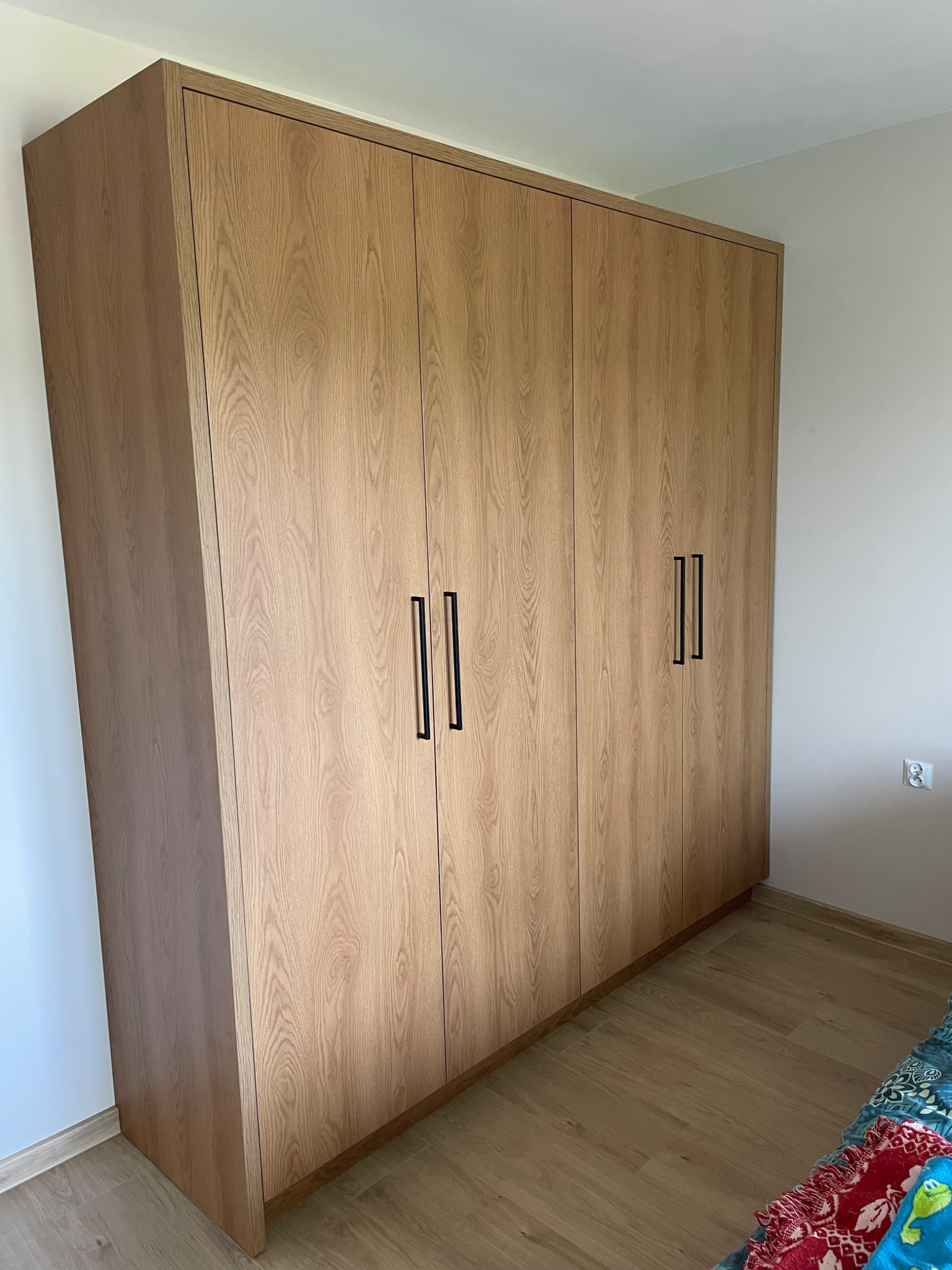 Duża, czterodrzwiowa szafa z jasnego drewna z czarnymi, pionowymi uchwytami, dopasowana do ściany w pokoju z drewnianą podłogą.