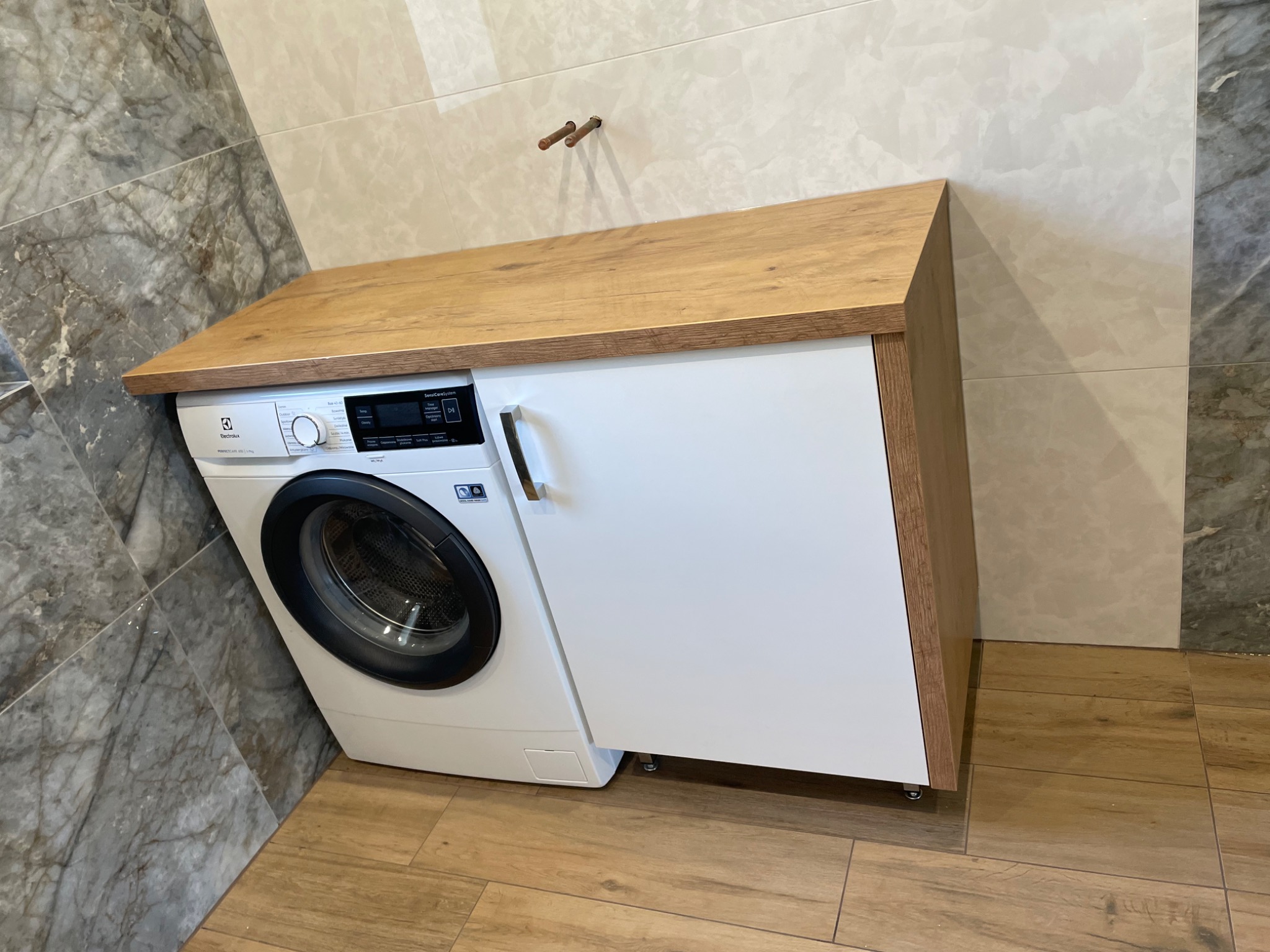 Pralka Electrolux zabudowana w szafce z drewnianym blatem w łazience wyłożonej płytkami imitującymi marmur i drewno.