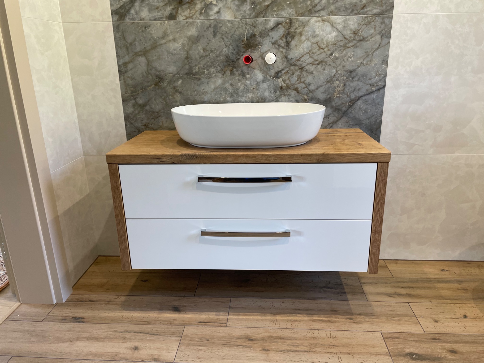 Nowoczesna łazienka z drewnopodobną podłogą i wiszącą szafką z blatem drewnianym, na której stoi owalna, biała umywalka nablatowa; na ścianie widoczne przyłącza wodne.