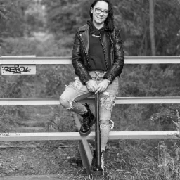 Czarno-białe zdjęcie kobiety w okularach, skórzanej kurtce i podartych dżinsach, opierającej się o metalową barierkę z graffiti, trzymającej kij bejsbolowy.