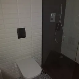 Nowoczesna łazienka z białą, podłużną glazurą, czarną armaturą i kabiną prysznicową typu walk-in z czarnym brodzikiem, widoczne wzorzyste płytki podłogowe.