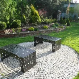 Dwie metalowe ławki z ażurowym wzorem liści na okrągłym brukowanym patio, otoczone zielonym trawnikiem i roślinnością ogrodową w tle.