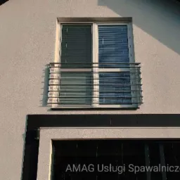 Nierdzewna balustrada balkonowa z poziomych rurek, zamontowana przed oknem z żaluzjami w jasnym budynku. Widoczny cień rzucany przez balustradę na ścianę.