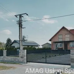 Nowoczesna brama wjazdowa z poziomymi, czarnymi przęsłami, osadzona na słupkach z szarej kostki, na tle domów jednorodzinnych i słupa energetycznego.