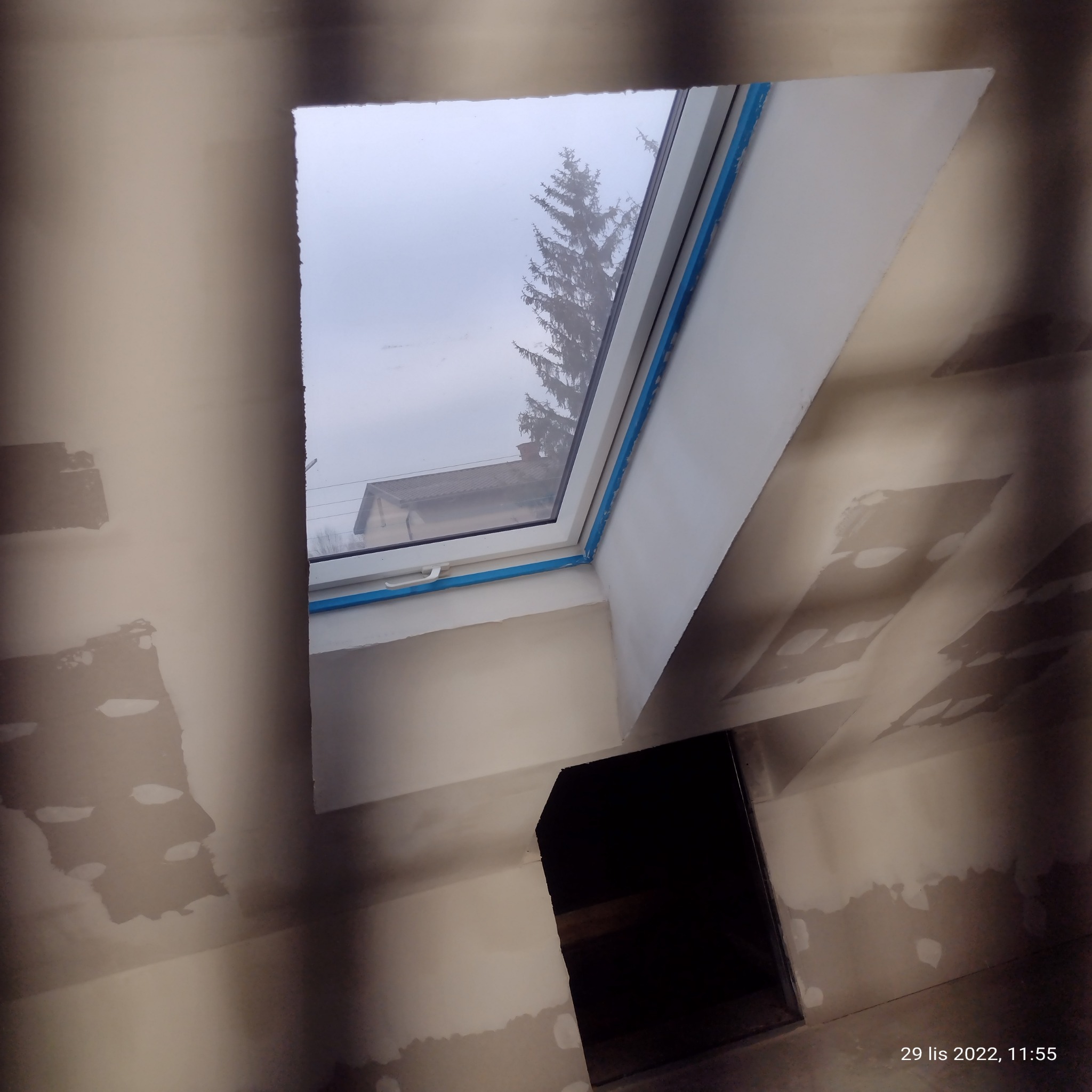 Wnętrze w trakcie prac wykończeniowych: zamontowane okno dachowe z niebieską taśmą zabezpieczającą, ściany ze śladami gładzi, widoczny otwór w ścianie.