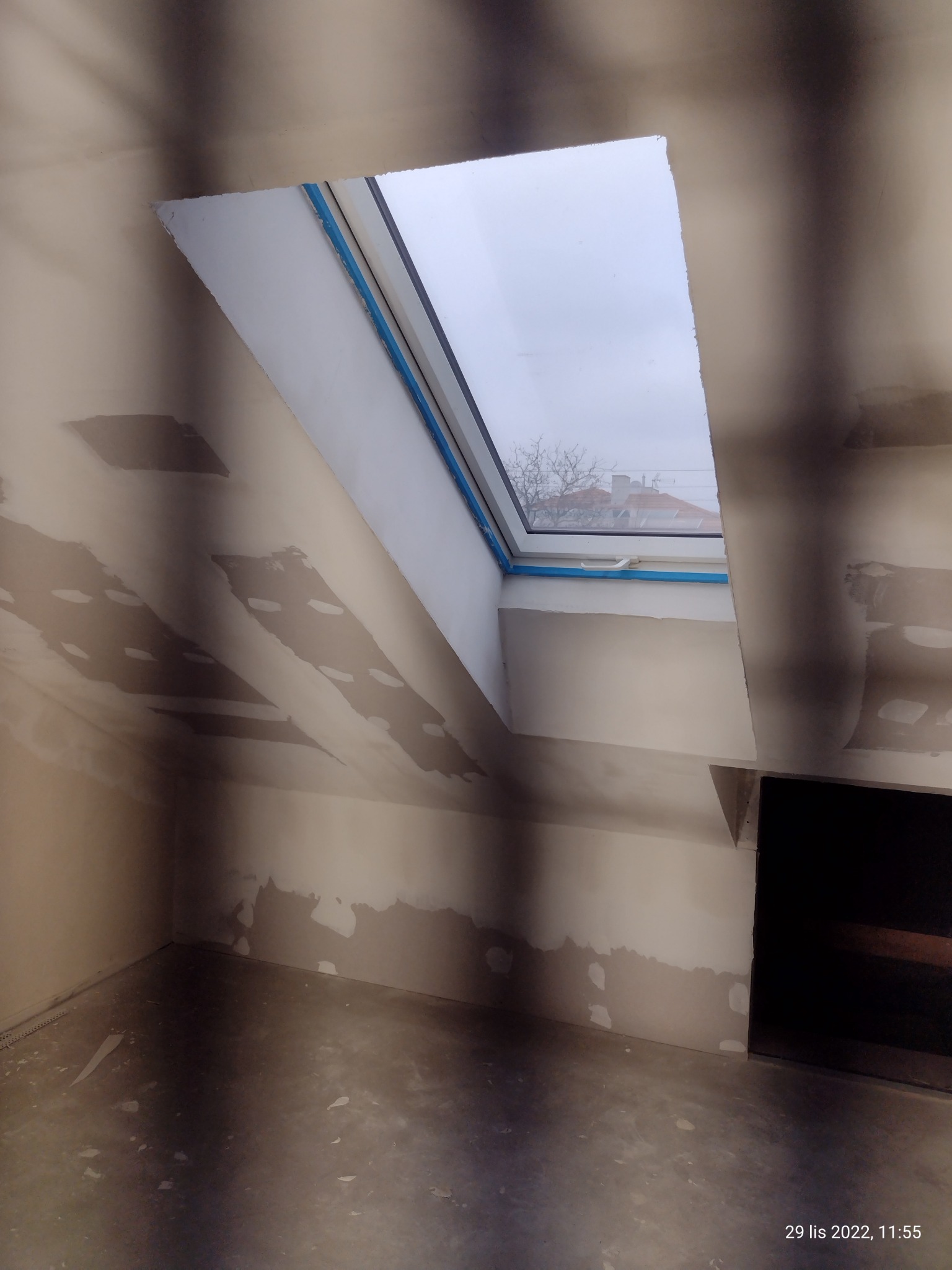 Poddasze w trakcie wykańczania: widok na okno dachowe z widokiem na dach sąsiedniego budynku, ściany częściowo pokryte gładzią, surowy beton na podłodze.