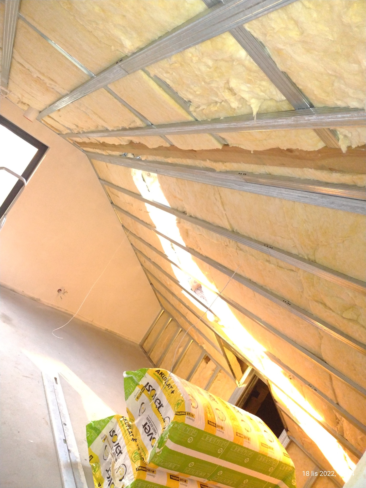 Poddasze w trakcie ocieplania wełną mineralną, widoczne metalowe profile konstrukcyjne i zapakowane paczki wełny Isover Akunat+ Thermi Star na podłodze.