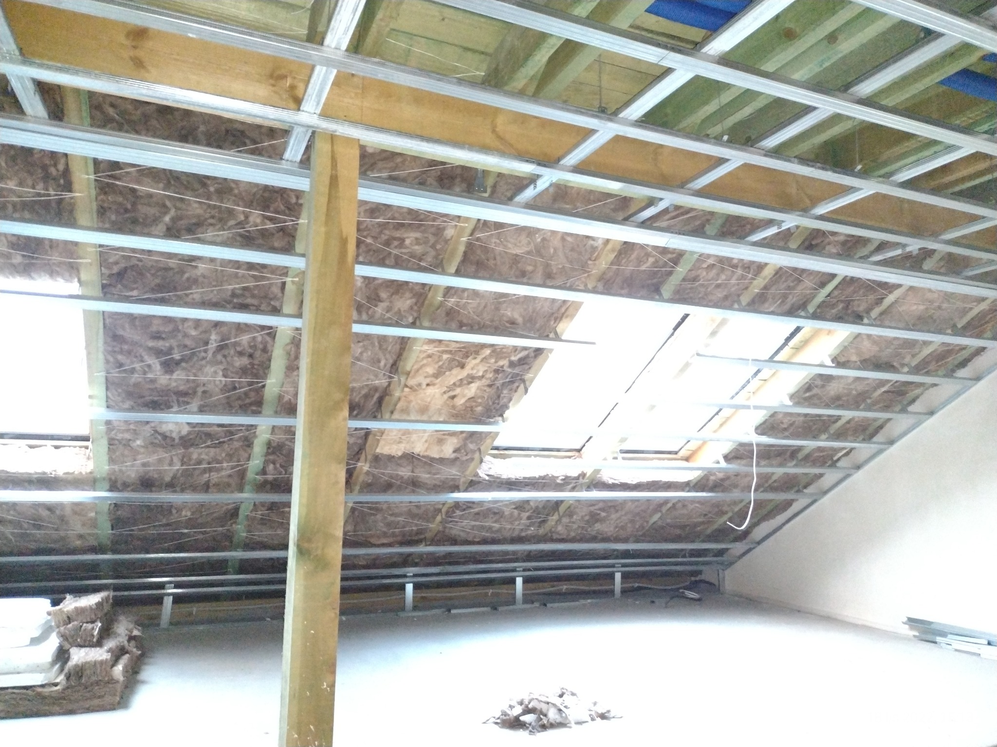 Skośny sufit poddasza w trakcie izolacji wełną mineralną, widoczne metalowe profile konstrukcyjne i drewniane elementy, na podłodze resztki materiałów izolacyjnych.