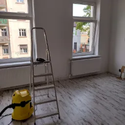 Pokój po remoncie: żółty odkurzacz Karcher i aluminiowa drabina stoją na podłodze z paneli imitujących drewno, dwa okna z widokiem na budynki.
