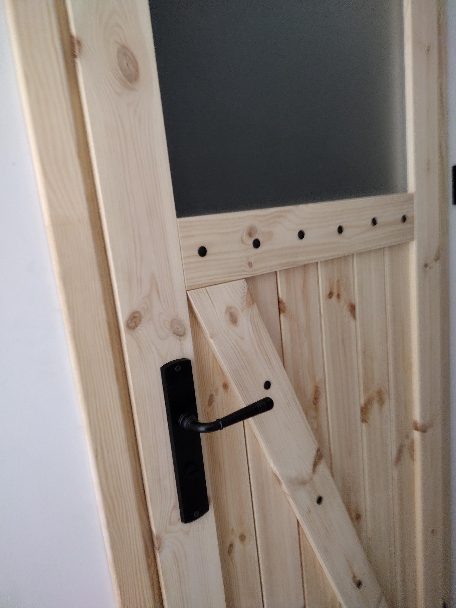 Jasne drewniane drzwi z czarną klamką i przeszkleniem, widoczne słoje drewna i czarne nity.