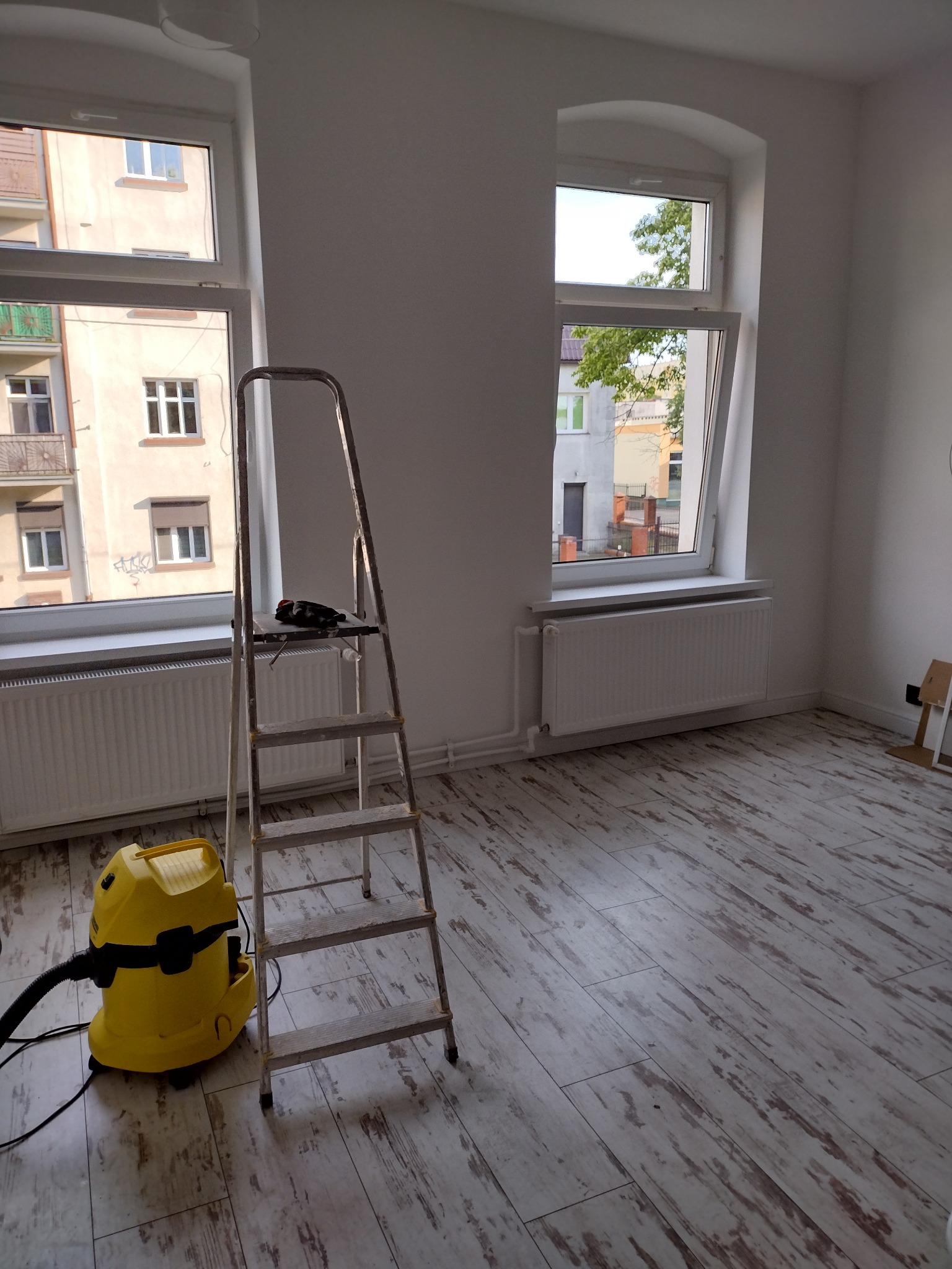 Pokój po remoncie: żółty odkurzacz Karcher i aluminiowa drabina stoją na podłodze z paneli imitujących drewno, dwa okna z widokiem na budynki.