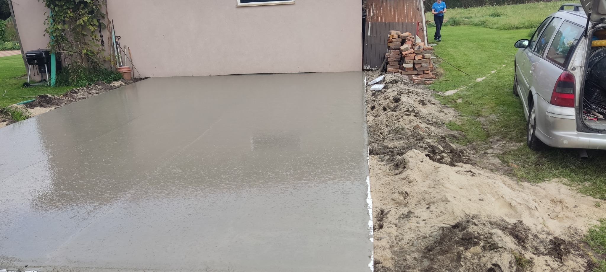 Świeżo wylana, gładka betonowa posadzka przed budynkiem, z widocznym stosem cegieł i samochodem z otwartym bagażnikiem w tle.