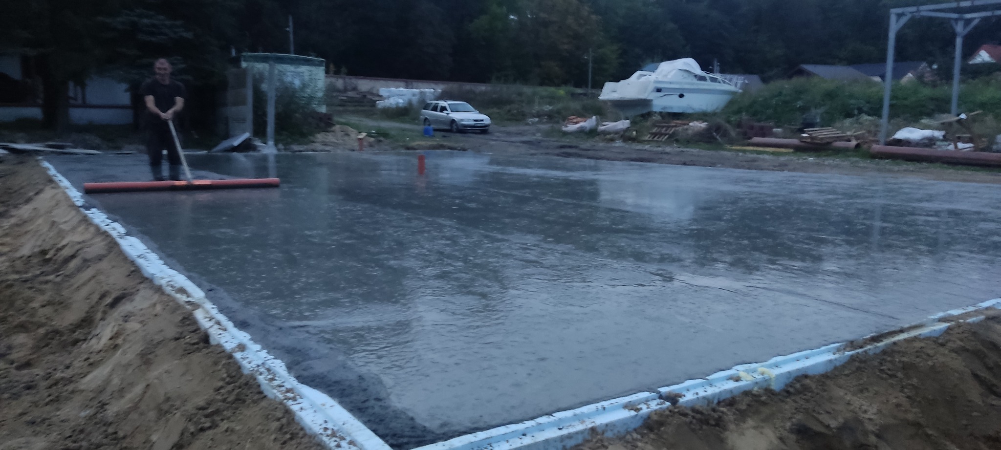 Wylewka fundamentowa z wygładzonym betonem, obramowana styropianem, w tle zaparkowany samochód i łódź pod plandeką.