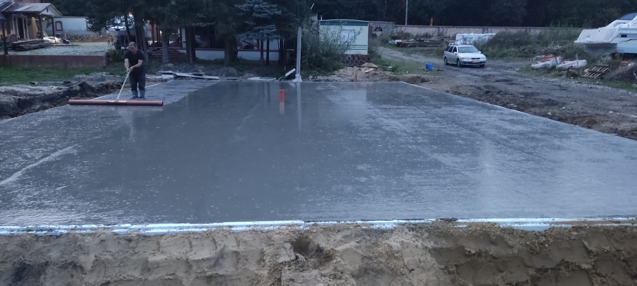 Wyrównywanie wylewki betonowej na przygotowanym podłożu, widoczny pracownik w kaloszach i z narzędziem do rozprowadzania betonu, w tle samochód osobowy i przyczepa kempingowa.