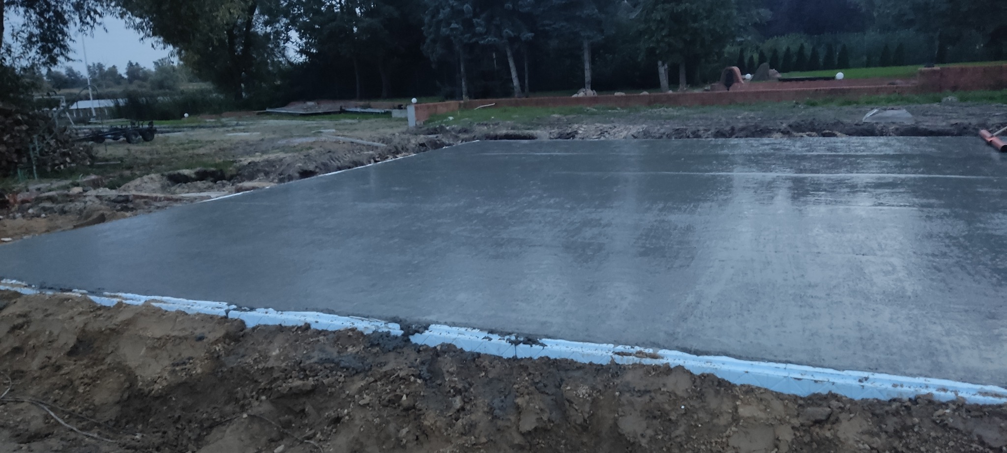 Świeżo wylana płyta fundamentowa z widocznym obrysem izolacji termicznej w gruncie, w tle drzewa i elementy krajobrazu.