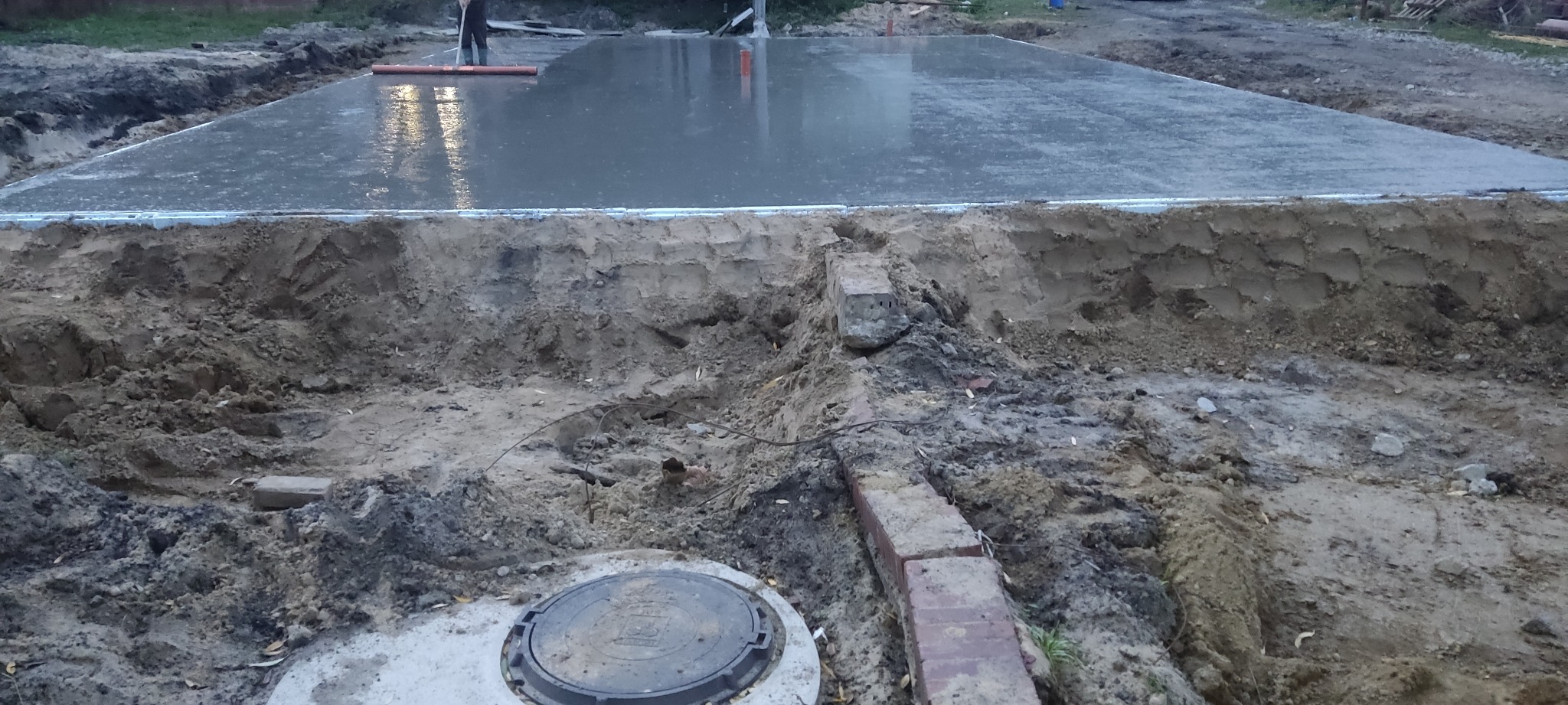 Wylewka betonowa fundamentu domu jednorodzinnego w trakcie wyrównywania, widoczne szalunki i wykop z ziemią, w tle osoba zacierająca beton.