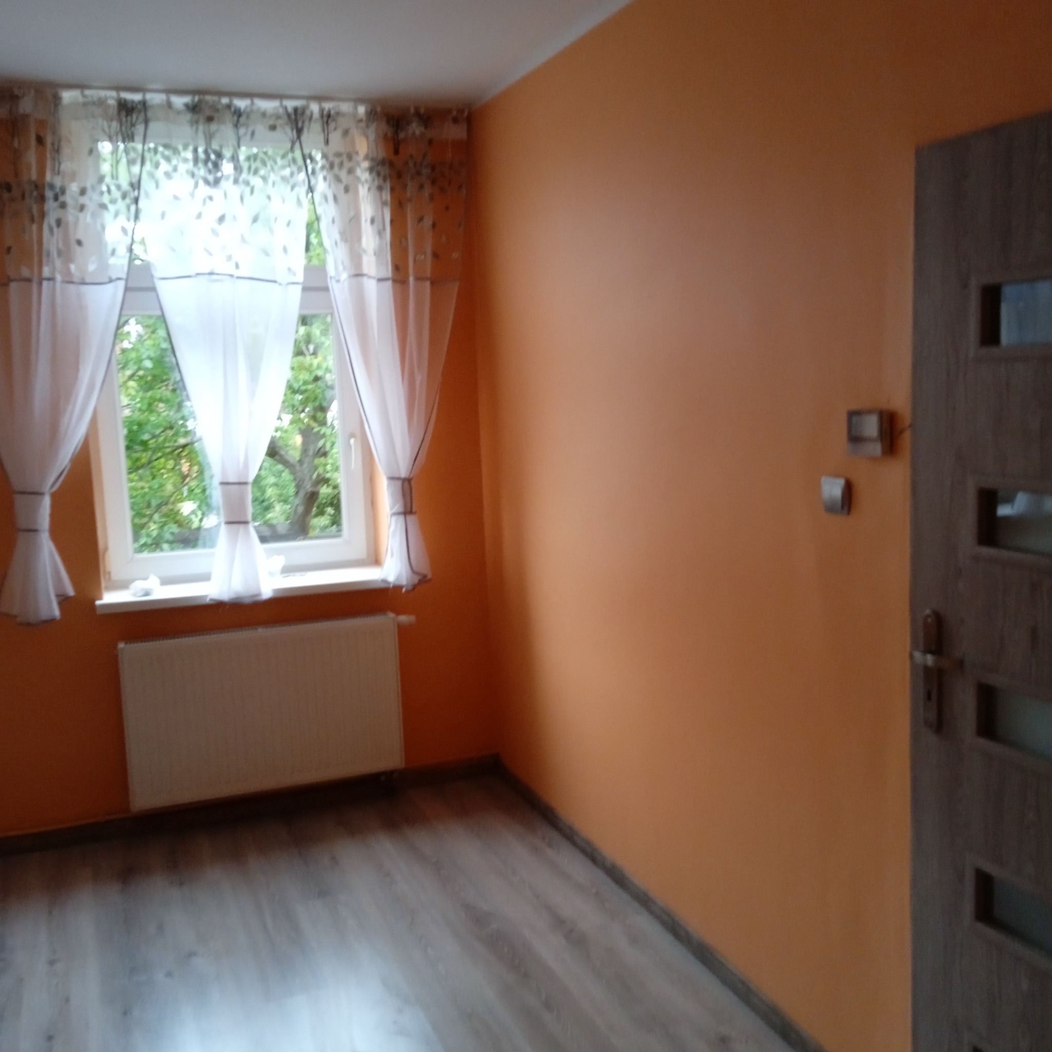 Pomarańczowy pokój z panelami podłogowymi, oknem z białymi firankami w drzewka i szarą, nowoczesną stolarką drzwiową.