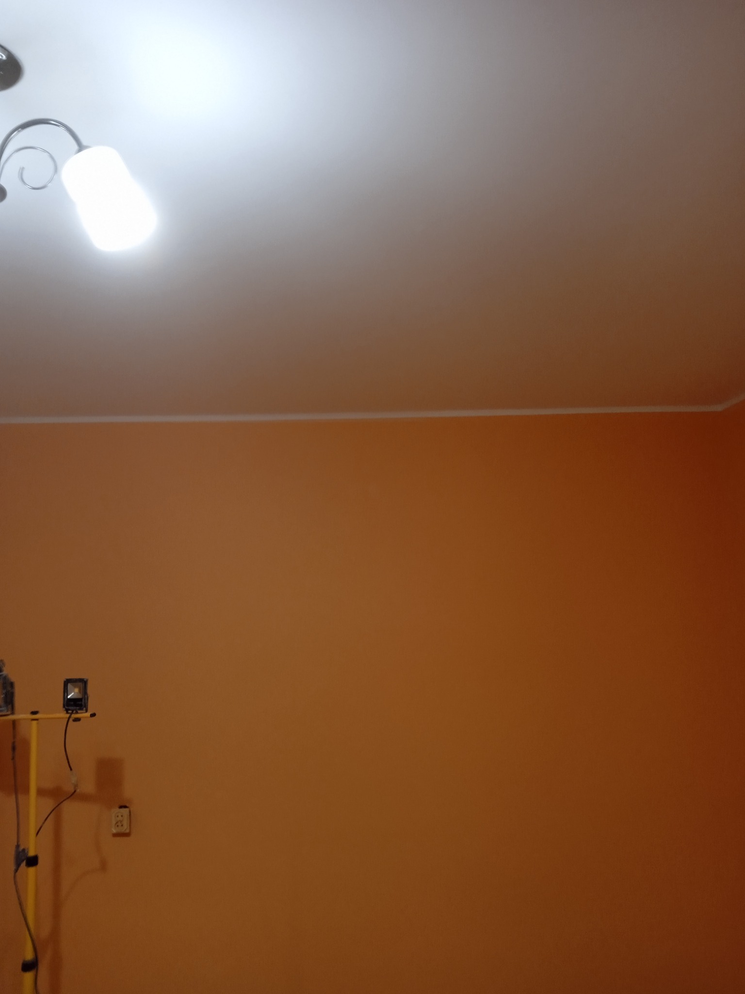 Pomarańczowa ściana po malowaniu, widoczna lampa budowlana na statywie i gniazdko elektryczne