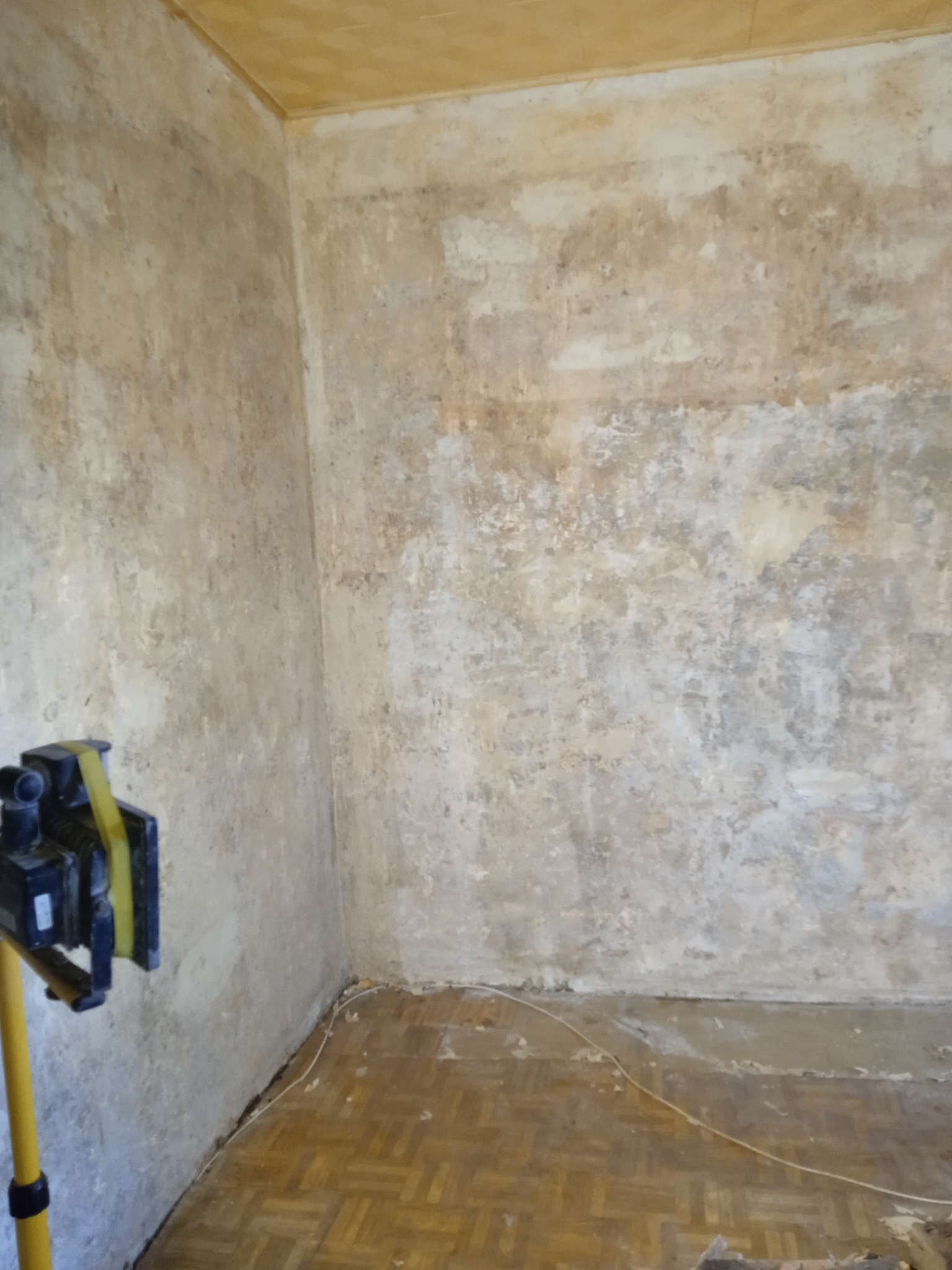 Pokój w trakcie przygotowania do remontu: zniszczone ściany z resztkami starej farby lub tapety, widoczna lampa budowlana i fragment parkietu z resztkami gruzu.