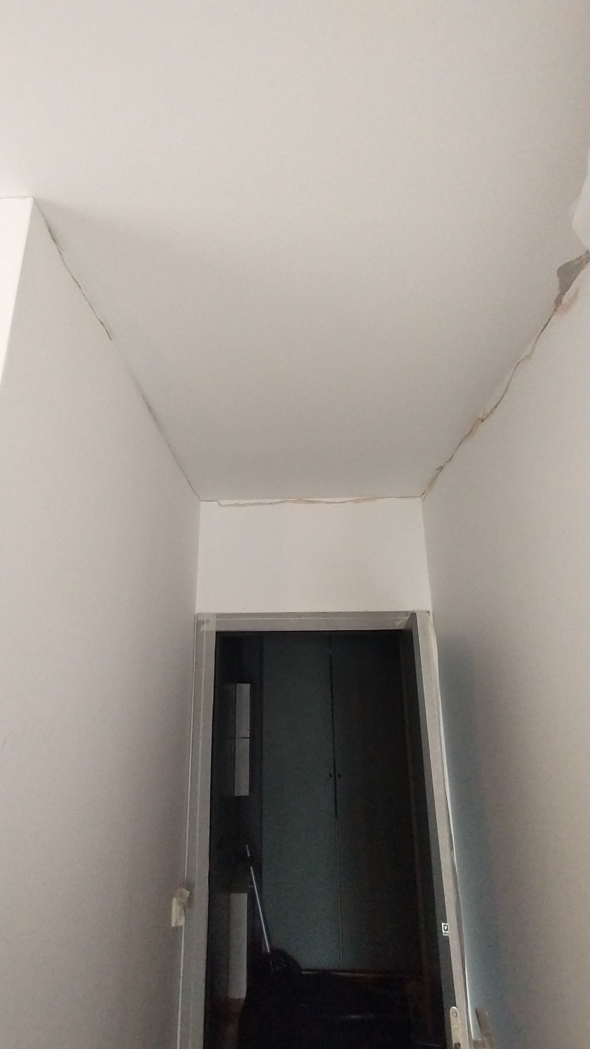 Biały sufit i ściany z widocznymi pęknięciami i uszkodzeniami tynku w rogu pomieszczenia, nad framugą drzwi.