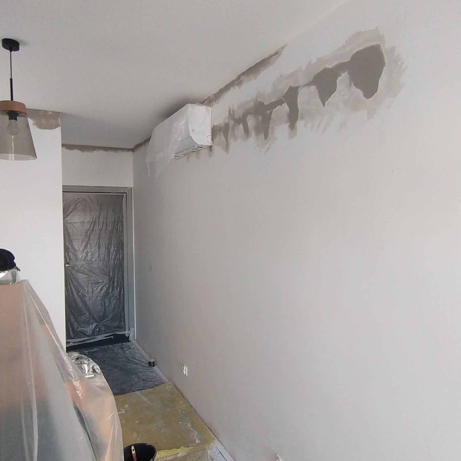 Przygotowanie ściany do malowania: załatana ściana z widocznymi śladami szpachli, klimatyzator zabezpieczony folią, podłoga zakryta folią ochronną, widoczna lampa i drzwi zabezpieczone folią.