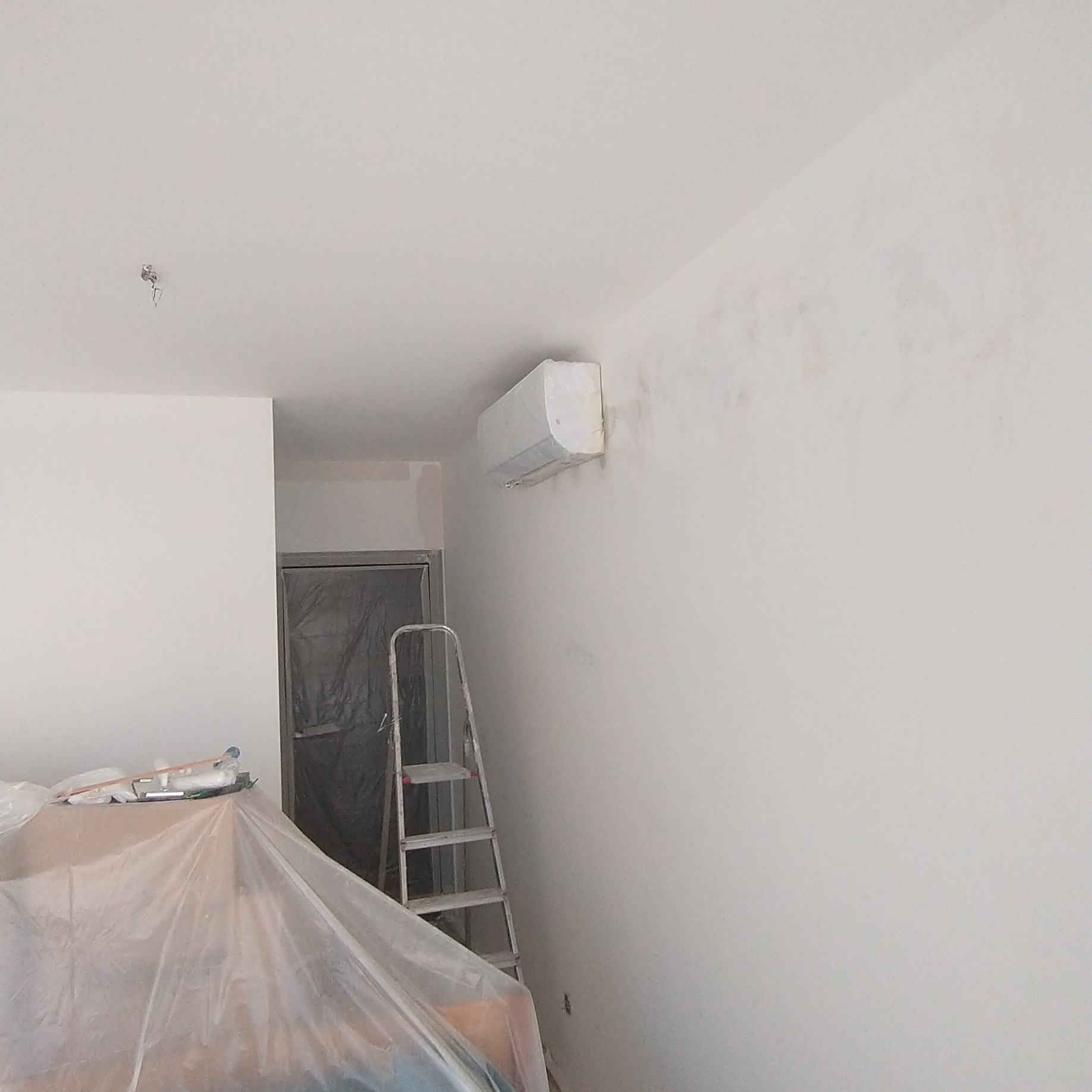 Pokój w trakcie odświeżania: drabina oparta o ścianę, klimatyzator zabezpieczony folią malarską, widoczne ślady po wcześniejszym malowaniu na ścianie, meble zakryte folią ochronną.