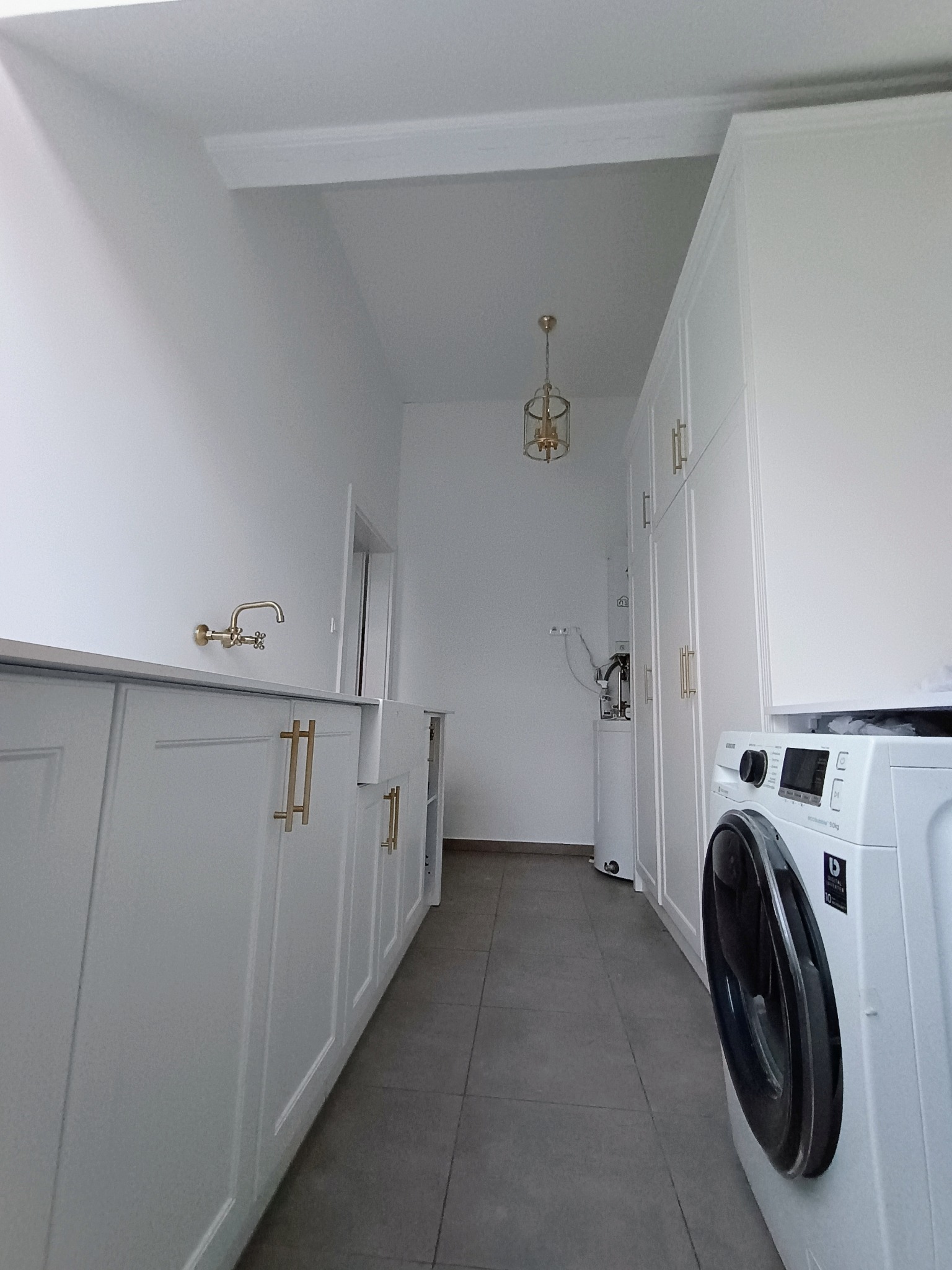 Biała pralnia z zabudową meblową: szafki z złotymi uchwytami, pralka Samsung i dekoracyjna lampa sufitowa.