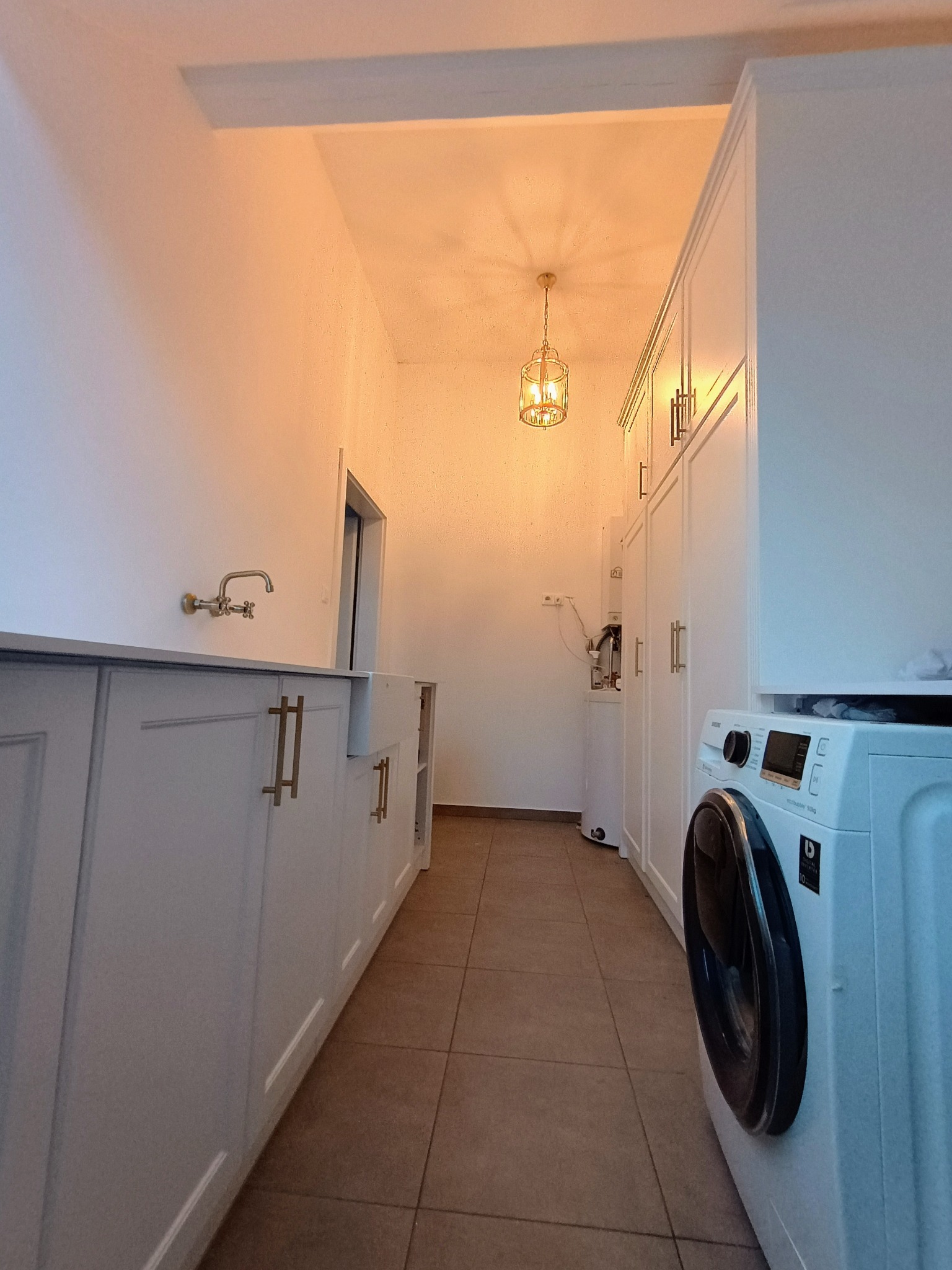 Wąskie pomieszczenie gospodarcze z białymi szafkami z frezowanymi frontami i złotymi uchwytami, ceramiczny zlew z baterią retro, pralka Samsung i dekoracyjna lampa wisząca.