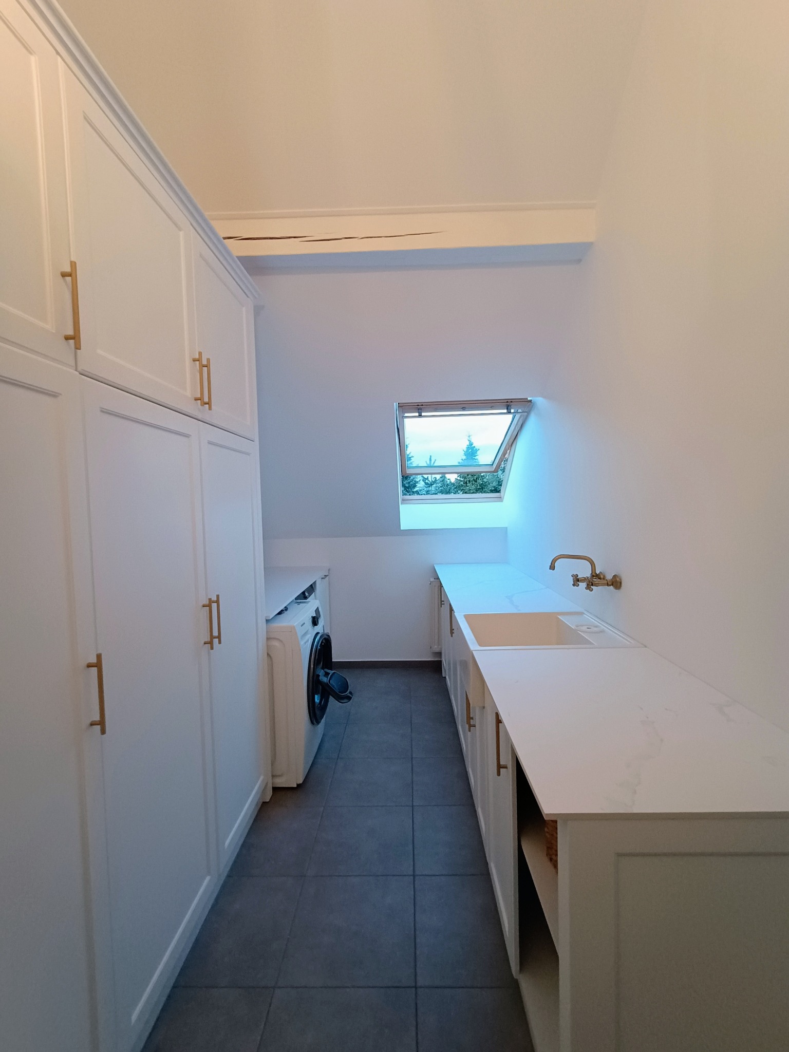 Biała pralnia na poddaszu z oknem dachowym, zabudową meblową z szafkami i blatem z umywalką, oraz pralką.