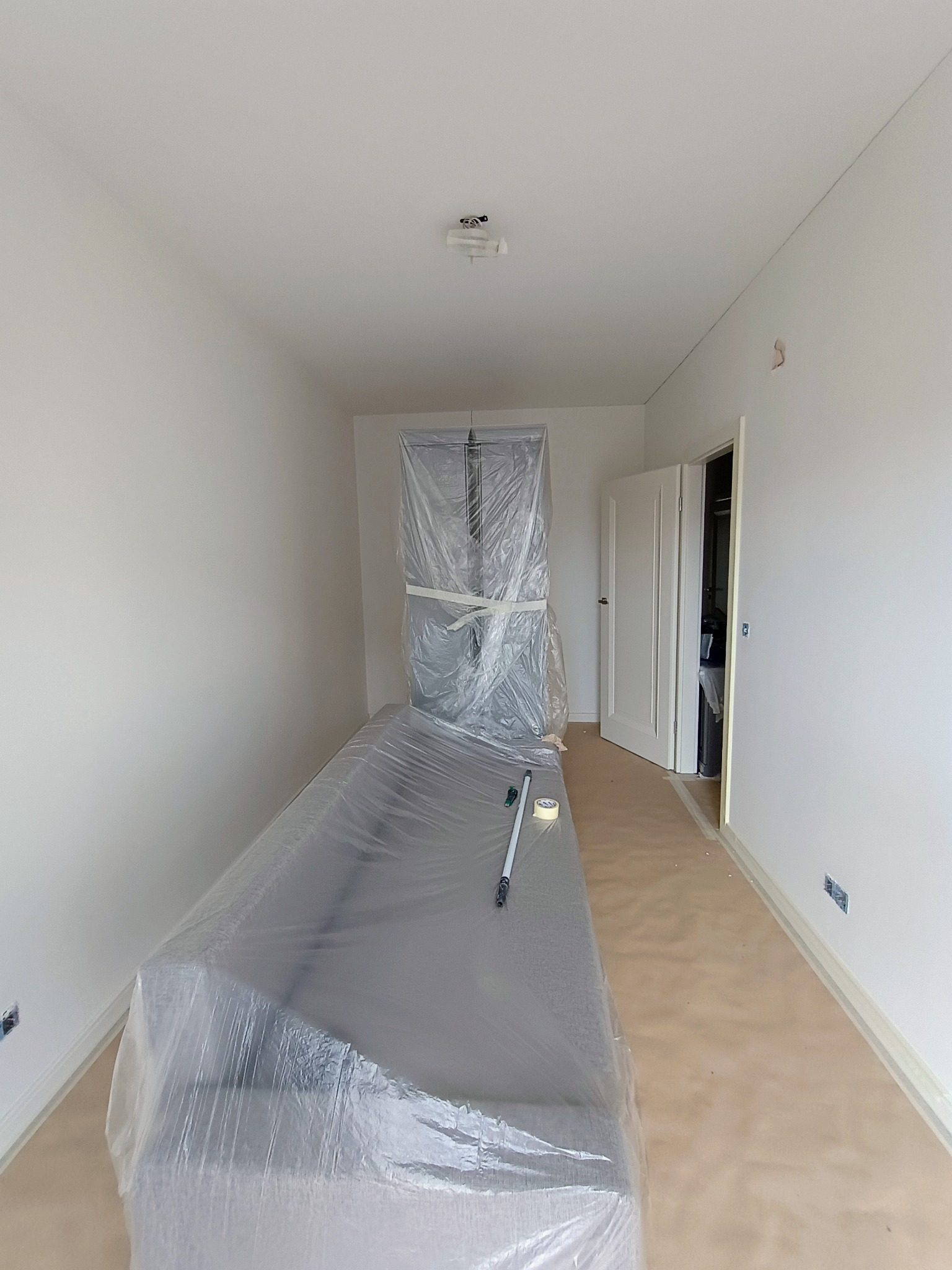 Pokój przygotowany do malowania: meble zabezpieczone folią, ściany białe, podłoga zaklejona taśmą malarską, widoczny wałek i taśma klejąca.
