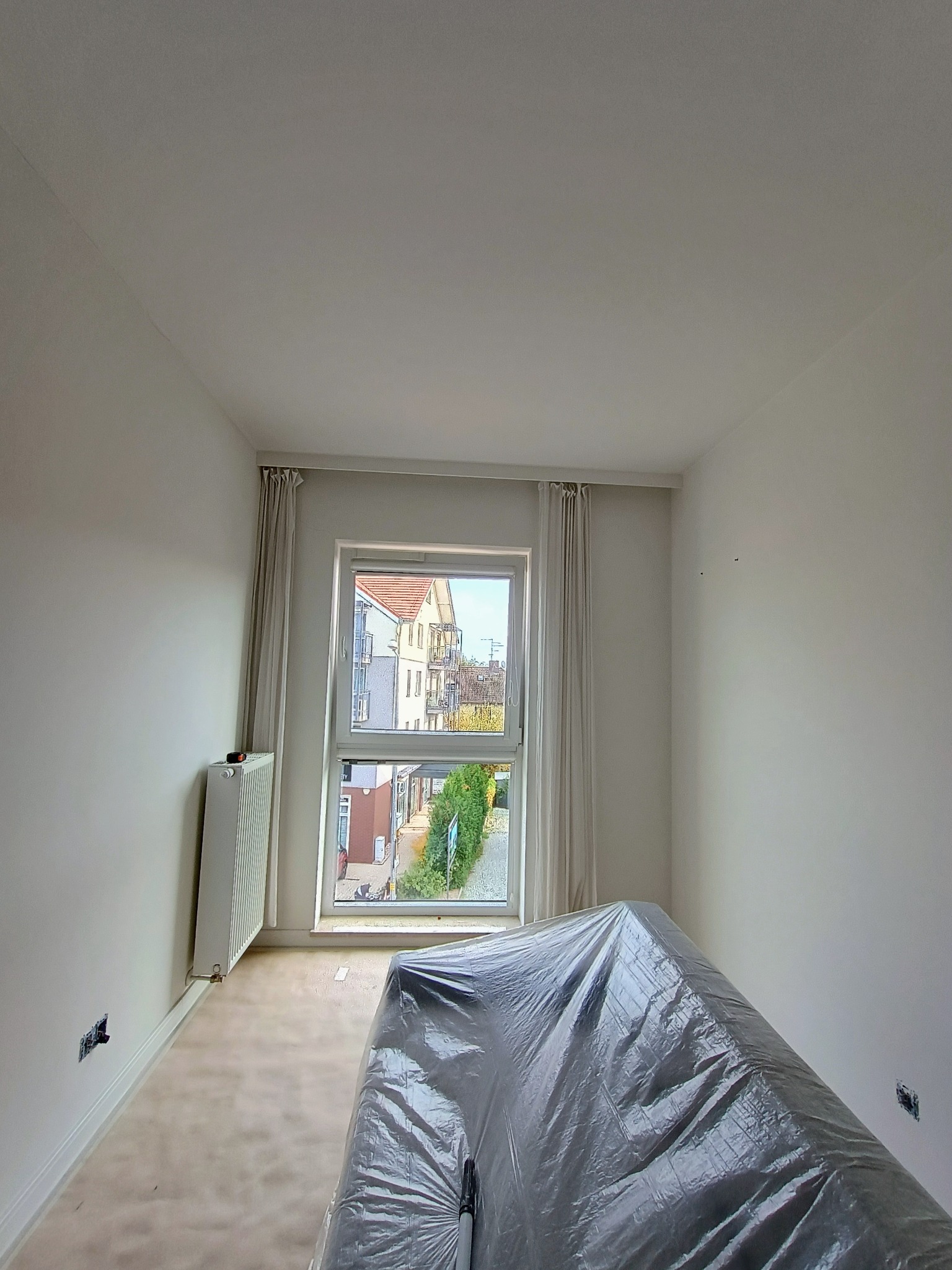 Pokój przed malowaniem: zabezpieczone meble folią, świeżo zagruntowane białe ściany, grzejnik, okno z widokiem na budynki.
