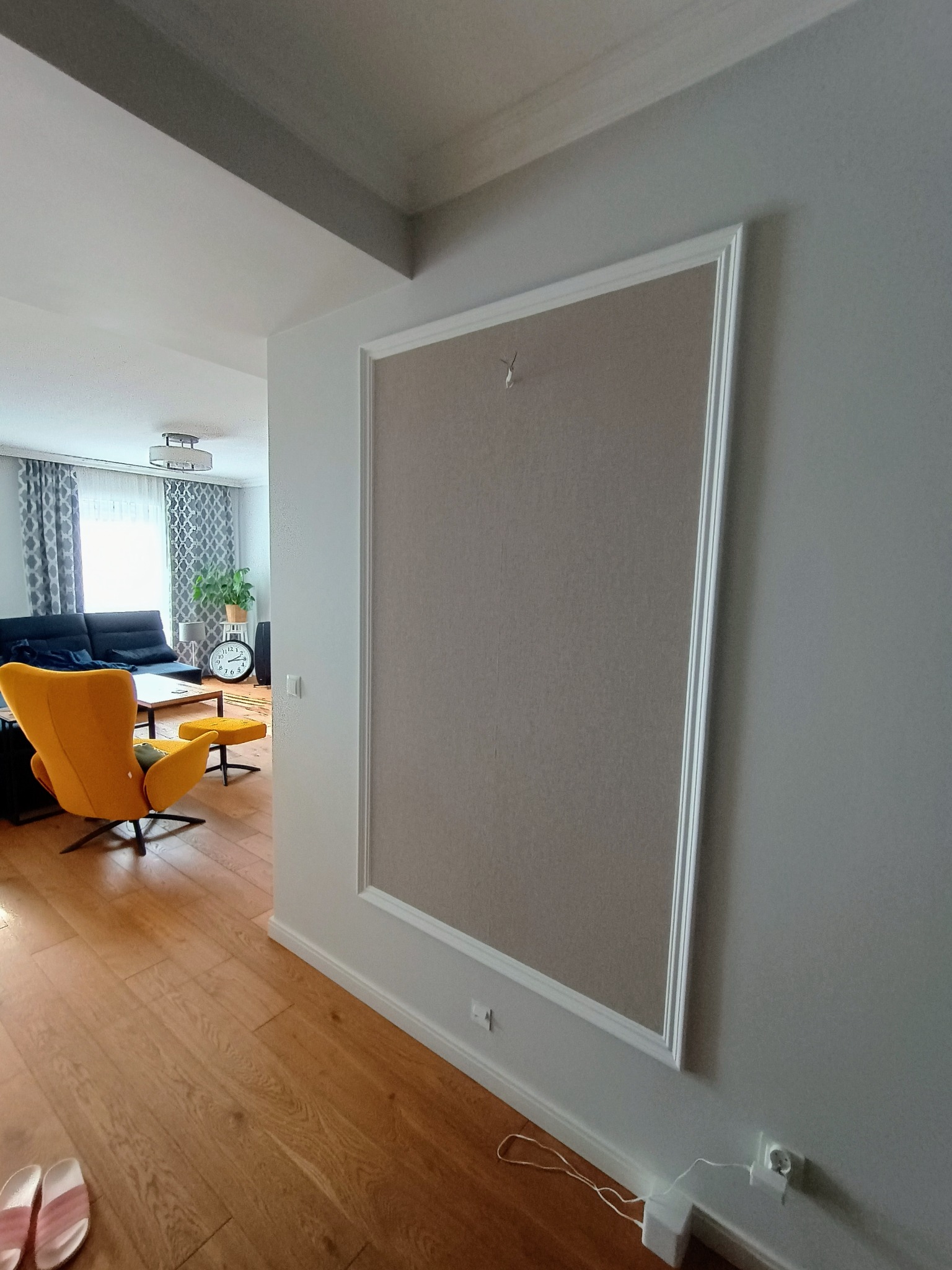 Szara ściana z dekoracyjną ramą w jasnym odcieniu, widok na salon z żółtym fotelem i sofą w tle.