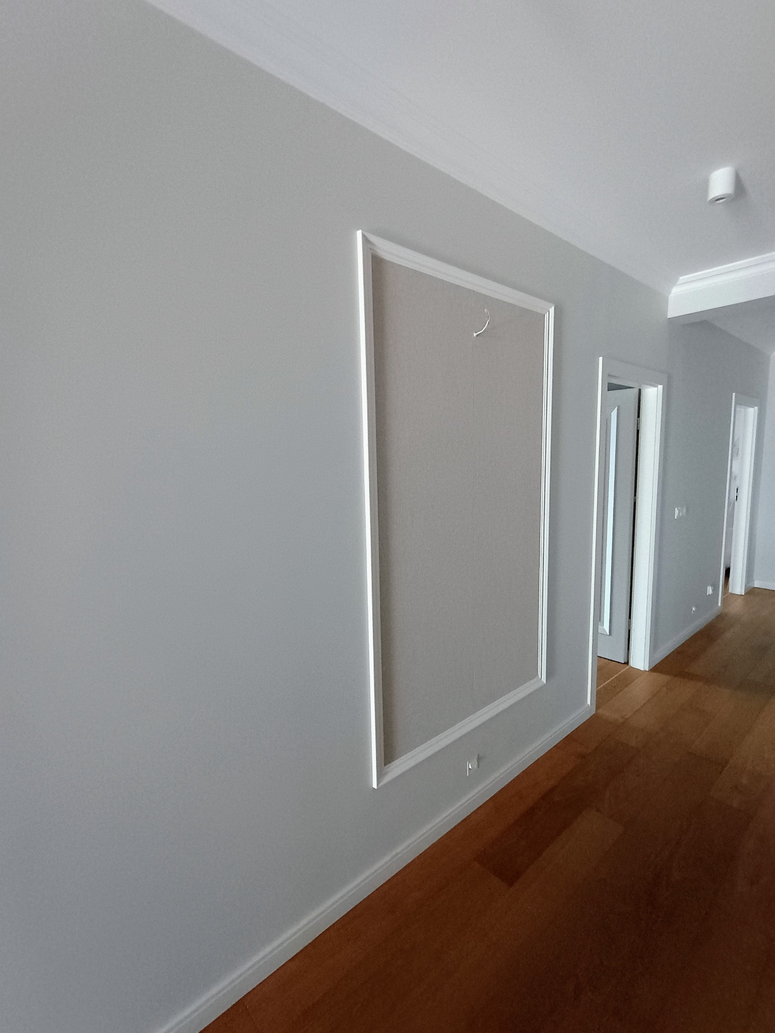 Szara ściana w korytarzu po remoncie, z drewnianą podłogą i białymi listwami przypodłogowymi, ozdobne obramowanie na ścianie z ukrytym włazem rewizyjnym.