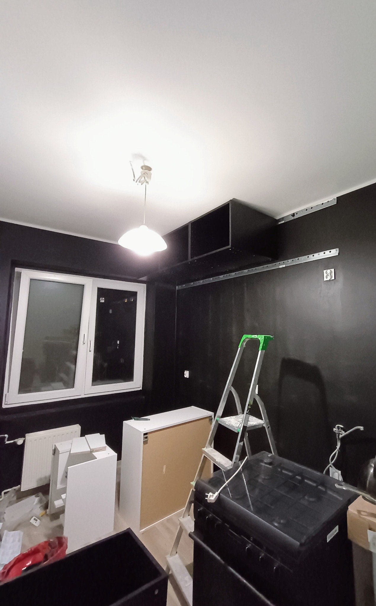 Pokój z czarnymi ścianami w trakcie montażu mebli: widoczna drabina, niezmontowane białe szafki, listwy montażowe na ścianie i wiszące szafki pod sufitem.