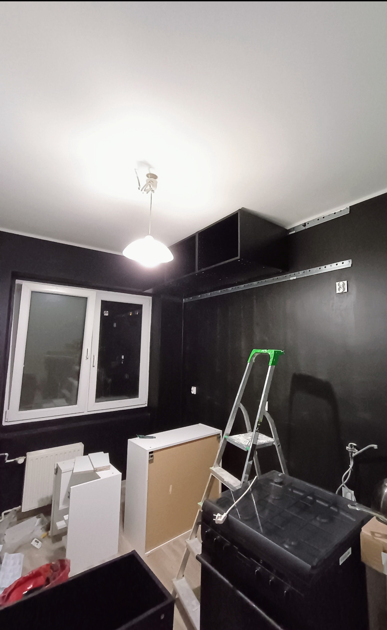 Pokój w trakcie remontu z czarnymi ścianami, widoczne niedokończone meble, drabina i lampa sufitowa, elementy montażowe na ścianie.