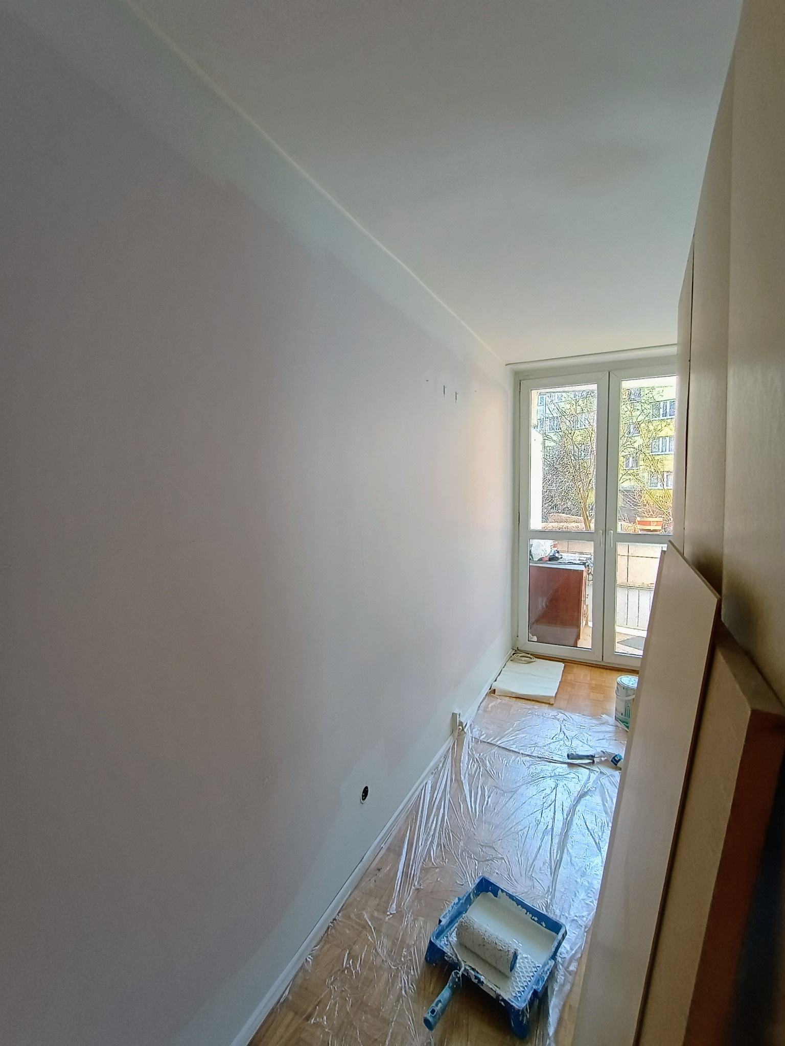 Pomieszczenie w trakcie malowania na jasny kolor: ściany częściowo pokryte farbą, podłoga zabezpieczona folią malarską, widoczny kuweta z wałkiem malarskim, okno balkonowe z widokiem na zieleń.