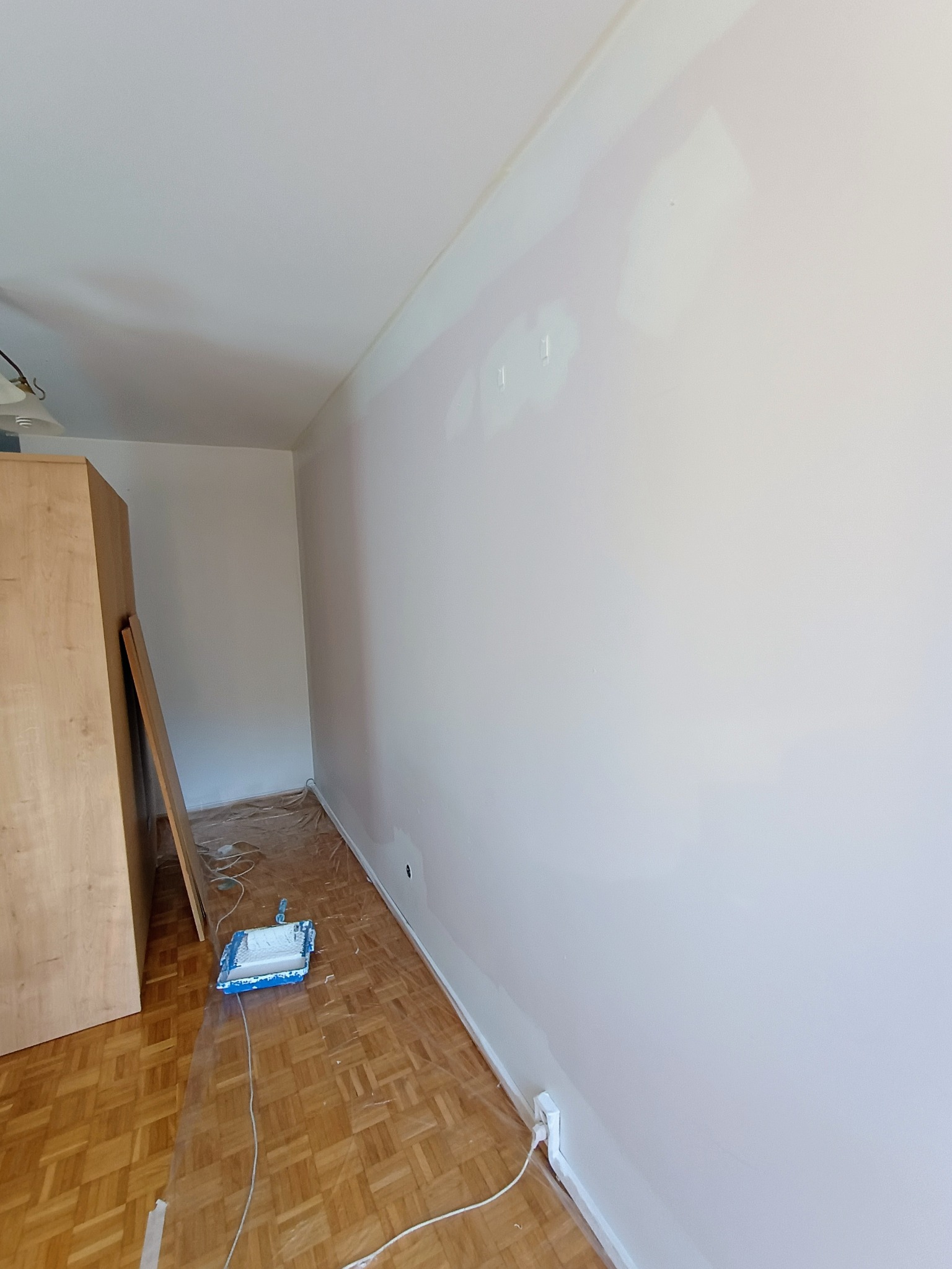 Pokój w trakcie odświeżania: ściana po nałożeniu pierwszej warstwy farby, widoczne niedociągnięcia, podłoga zabezpieczona folią malarską, kuweta z wałkiem na podłodze, drewniana szafa z boku.