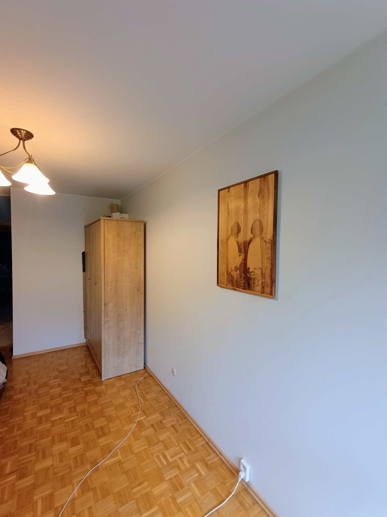 Jasne wnętrze pokoju z parkietem, szafą i obrazem na ścianie pomalowanej na jasny odcień niebieskiego.