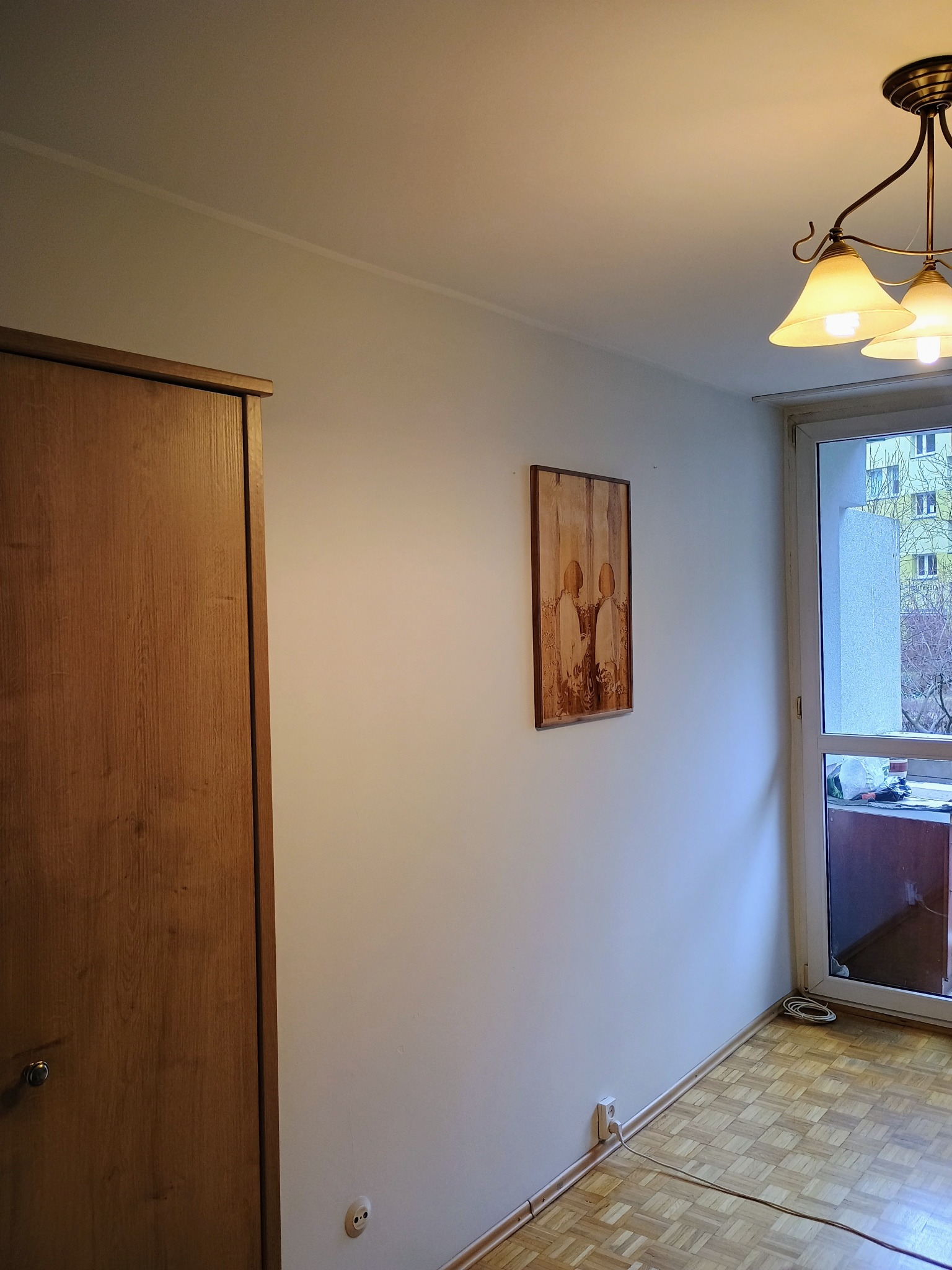 Wnętrze pokoju z drewnianą szafą, obrazem na ścianie i oknem balkonowym, oświetlone lampą sufitową. Widoczny parkiet na podłodze.