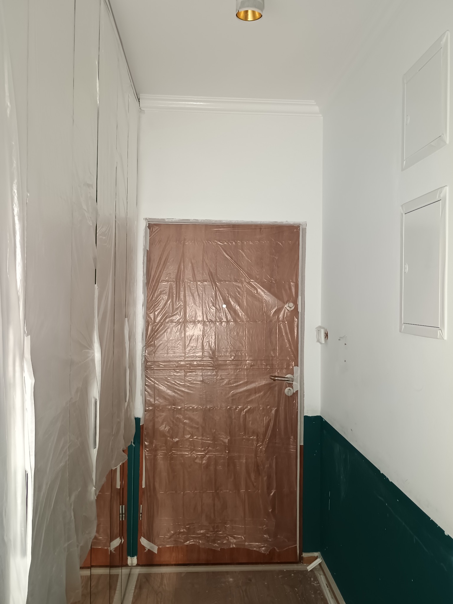 Korytarz w trakcie malowania: ściany częściowo pomalowane na biało i zielono, drzwi i szafa zabezpieczone folią malarską. Widoczne niedociągnięcia i przygotowania do dalszych prac.