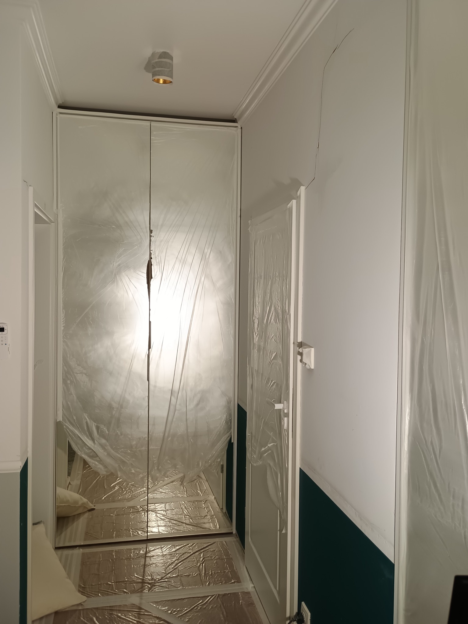 Przygotowanie do malowania: folią zabezpieczone meble, podłoga i drzwi w korytarzu z widoczną pękniętą ścianą i pomalowaną dolną częścią ścian na ciemnozielono.