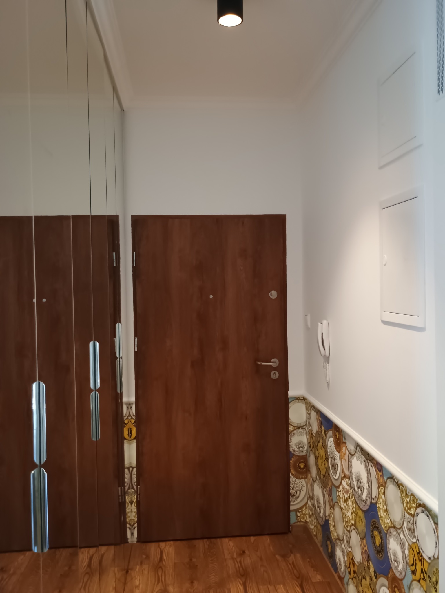 Korytarz po remoncie: drewniane drzwi wejściowe, szafa z lustrami, dekoracyjne płytki imitujące talerze wzdłuż ściany i drewnopodobna podłoga. Białe ściany i sufit.