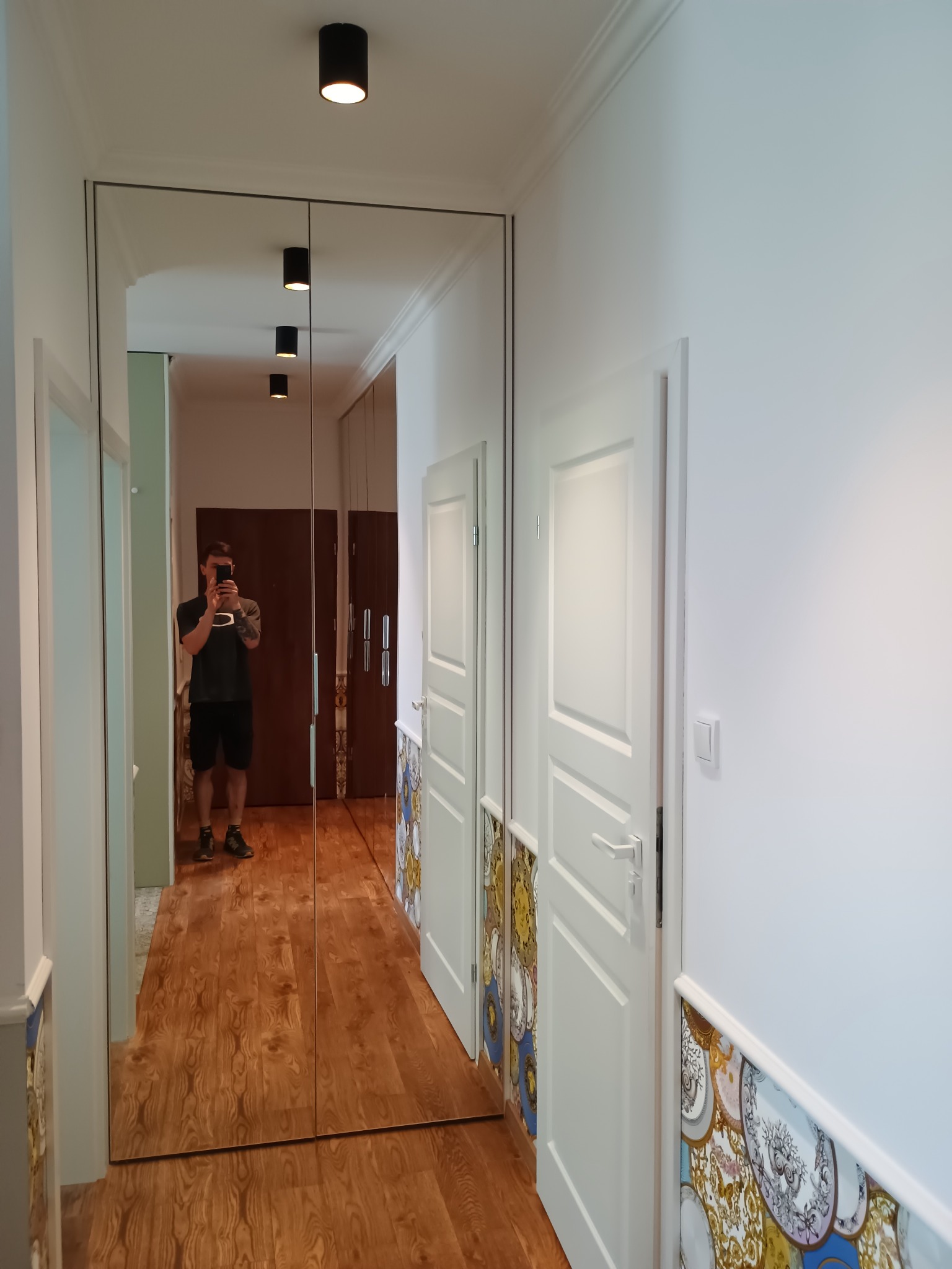 Korytarz po remoncie z lustrzaną szafą wnękową i odnowionymi drzwiami z dekoracyjnym malowaniem paneli. Widoczne oświetlenie sufitowe i podłoga imitująca drewno.